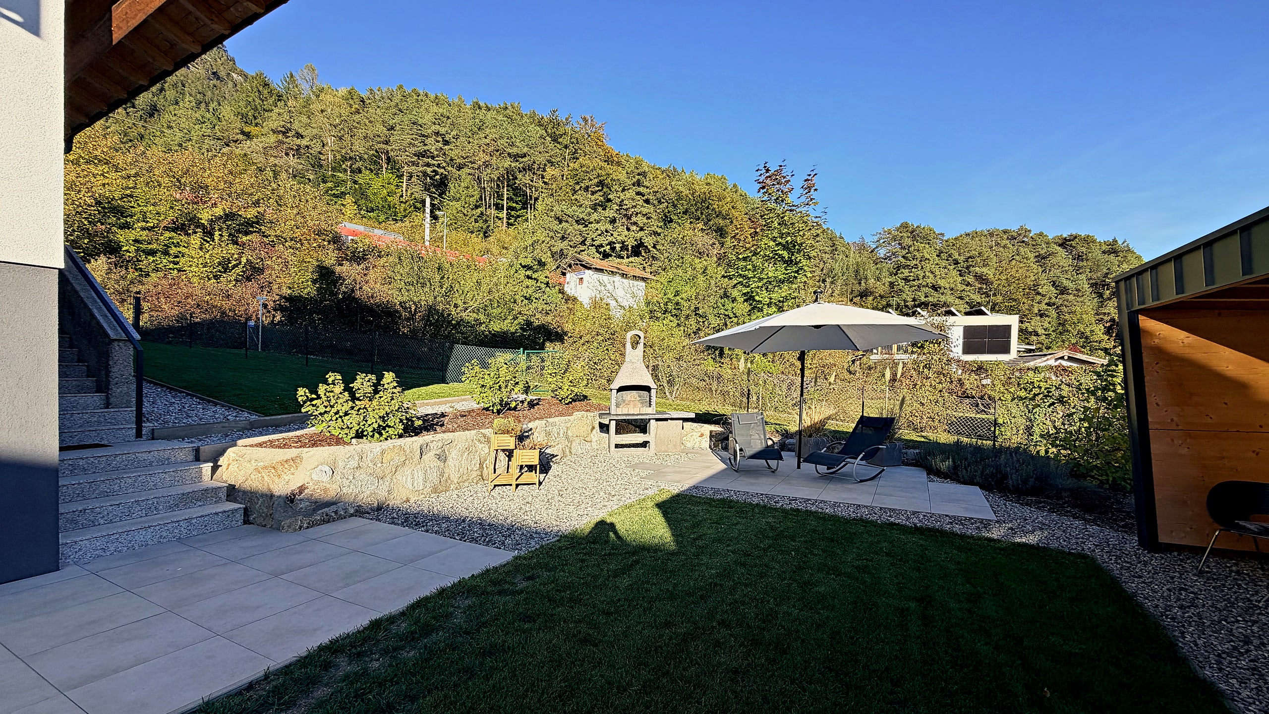Well-kept lawn with lounging area, parasol and built-in barbecue in front of a wooded slope